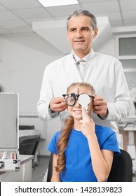 Front View Of Happy Girl Wearing Glasses With Lens For Checking Vision. Petty Child Closing Eye With Hand And Looking At Letters While Optician Standing Behind In Clinic. Concept Of Eye Care.