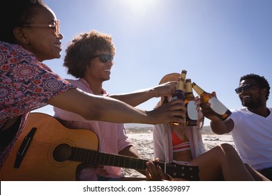 Front view of happy multi ethnic group of friends toasting beer bottle while sitting on the beach in the sunshine