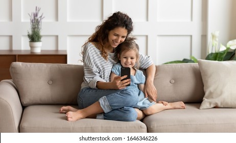 Front View Happy Pretty Mommy Cuddling Small Kid Daughter, Taking Selfie Shot On Smartphone Or Playing Online Game At Home. Loving Young Mother Embracing Little Child, Using Mobile Apps Together.