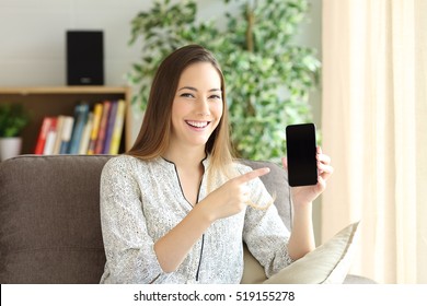 Front view of a happy woman showing a black phone screen on line and presenting a new product in a house interior