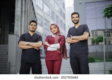 Front View Of High-skilled Team Of Three Young Confident Asian Arab Doctors, Standing Outside The Hospital And Looking At Camera. Pretty Muslim Lady In Hijab Stands Between Her Two Male Colleagues