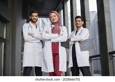 Front View Of High-skilled Team Of Three Young Confident Saudi Arab Doctors, Standing Outside The Hospital And Looking At Camera. Pretty Muslim Lady In Hijab Stands In Front Of Her Two Male Colleagues