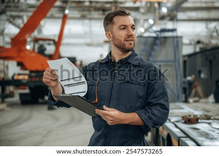 Front view, holding notepad, standing. Male worker is in the modern warehouse or manufacturing company.