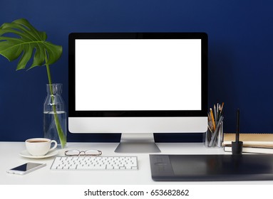 Front view of home office graphic designer desktop with blank copy space PC computer, graphic tablet, mobile phone, coffee cup, stationery and leaf Monstera on deep blue wall background. Mock up - Powered by Shutterstock