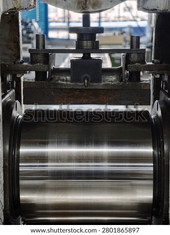 Front view of large industrial roller press showing polished rotating steel cylinder, heavy welded clamp assembly, textured adjustment knob and threaded bolt, worn metal surfaces and mounting bolts