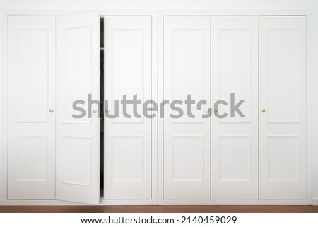 Front view of large white wardrobe in american classic style with ajar door in light room. Advertising of ergonomic furniture and spacious closet at modern apartment interior