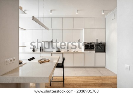 Front view of light minimalistic kitchen with white facade, built-in appliances and marble table. Stylish kitchenware on countertop. Well organized space in new apartment
