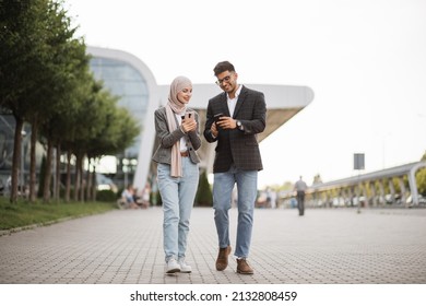 Front view of likable happy hindu man and muslim lady in hijab, walking on the street with smartphones, showing some posts or news in social networks and smiling - Powered by Shutterstock