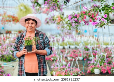Front view looking at camera florist aged woman in hat stands in greenhouse among indoor flowers in pots. Dressed in casual clothes. Smiles happily and holds favorite pot flower in hands. Copy space.