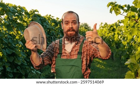 Front view looking at camera young farmer male winemaker bearded in the vineyard laughing with one hand holding his hat with the other thumb up. The winemaker is in his field in fine weather.