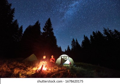 Front View Of Loving Couple Tourists Sitting By Bright Burning Campfire Near Two Tents, Enjoying Beautiful Camping Night Together Under Dark Sky Full Of Stars And Bright Milky Way, Warm Summer Night.