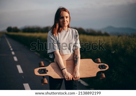 Front view , low angle .Funny young girl in jeans and top stands with big longboard. Green nature background