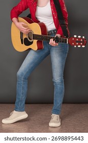 Front view of low section of unrecognizable woman guitarist playing six string acoustic guitar. Stylish young female musician is dressed in crimson leather jacket, jeans and sneakers