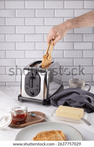 Front view of a male hand using wooden tongs to take hot toast from a chrome toaster. On marble countertop with butter, strawberry jam, cup and gray napkin. Copy space. 
