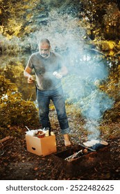 Front view of a man standing next to a campfire at autumn forest, he prepare food in the fire. The concept of tourism, camping, tourism.
