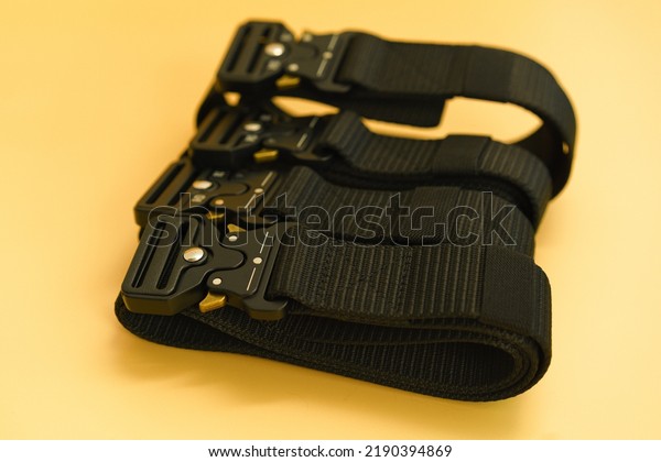 A front view of many black canvas belts with a\
black fast open metal male buckle and a black belt loop lays in the\
yellow background. Belt. Military. Buckle. Clothing. Equipment.\
Canvas. Protection