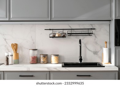 Front view of marble countertop with black sink near light ceramic wall and gray furniture in nordic style kitchen. Various jars with food and cooking utensils on stone surface.