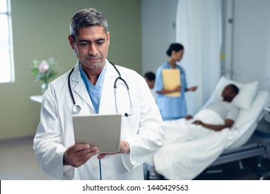 Front view of mature Caucasian male doctor using digital tablet while mixed race doctor discussing with African american man lying in bed in the ward at hospital