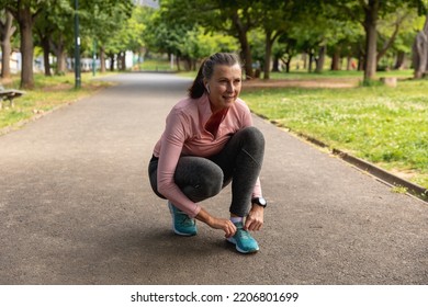 Front View Of A Mature Middle Aged Caucasian Woman Working Out In A Park, Squatting, Tying Her Shoe Laces On A Track