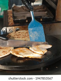 front view, medium distance of an open air, tropical, Greek vender is cutting grilled chicken on a flat iron grill to be sold to people waiting to be served 