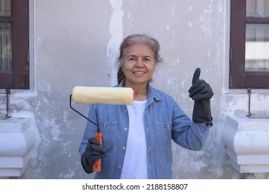 Front View Medium Shot Portrait Of Happy Senior Asian Woman Wearing Gloves, Holding Paint Roller, Smiling, Putting Thumb Up. Worker, Retiree Lifestyle, DIY Home Renovation Concept.