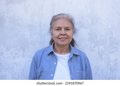 Front View, Medium Shot Portrait Of Gray Hair Senior Asian Woman Looking At Camera, Smiling, Wearing White T-shirt And Blue Shirt, In Front Of White Faded Old Wall. Retiree Lifestyle Concept.