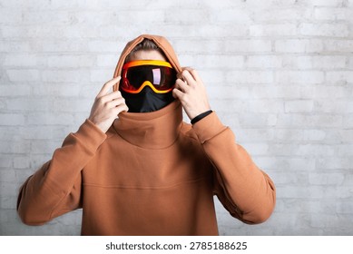 A front view medium studio shot of an unrecognizable winter sports enthusiast or model. The person is wearing a heavy brown (terracotta color) oversized hoodie with the hood pulled up and a black face balaclava mask covering the lower face. They are using both hands to adjust bright orange and red reflective ski or snowboard goggles. Set against a white brick wall background. Great for alpine gear retail, sportswear mockups, winter fashion, and extreme sports apparel.