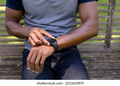 Front View Mid Section Of An African American Man With Gray T Shirt Sitting On A Bench In The Garden Checking His Smartwatch 