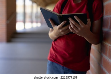 Front View Mid Section Of A Caucasian Schoolboy Wearing A Rucksack And A Red T Shirt Leaning Against A Wall In An Outdoor Corridor Reading A Book At Elementary School