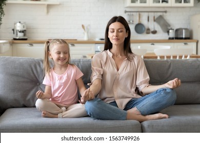 Front View Mindful Calm 30s Woman Sitting In Lotus Pose With Cute Little Daughter On Comfortable Sofa In Living Room. Smiling Preschool Girl Practicing Yoga Breathing Exercises With Mommy At Home.