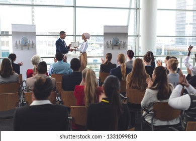 Front view of mixed race businesswoman receiving award from mixed race businessman in front of business people applauding at business seminar in office building
