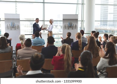 Front view of mixed race businesswoman receiving award from mixed race businessman in front of business professionals applauding at business seminar in office building - Powered by Shutterstock