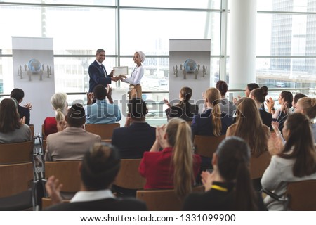 Front view of mixed race businesswoman receiving award from mixed race businessman in front of business professionals applauding at business seminar in office building