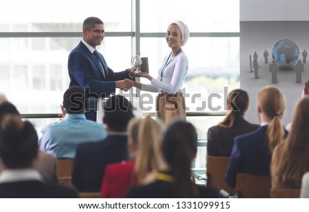 Front view of mixed race businesswoman receiving award from mixed race businessman in front of business professionals sitting at a business seminar in office building