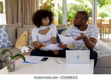 Front View Of A Mixed Race Couple Sitting In Their Living Room, Sitting On The Couch, Looking At Paperwork And Having A Discussion, A Laptop Computer And Smartphone On The Table 