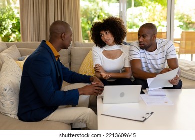 Front View Of A Mixed Race Couple Sitting In Their Living Room Talking With An African American Male Financial Adviser Using A Laptop Computer 