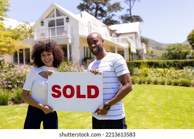 Front view of a mixed race couple standing in the garden, smiling to camera and holding sign with the word sold written on it  - Powered by Shutterstock