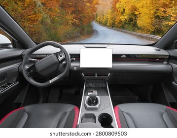 Front view of a modern car interior dashboard with blank white screen mockup facing a scenic autumn road trip view through the windshield. Luxury vehicle cockpit with center console on a beautiful seasonal highway drive.