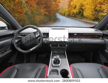 Front view of a modern car interior dashboard with blank white screen mockup facing a scenic autumn road trip view through the windshield. Luxury vehicle cockpit with center console on a beautiful seasonal highway drive.