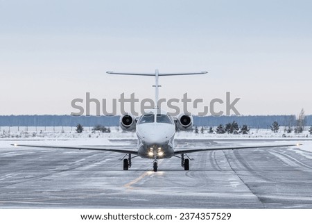 Front view of the modern white private jet taxiing on airport taxiway in winter