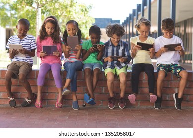 Front view of multi ethnic students using digital tablet while sitting on brick wall at corridor in school