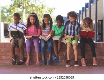 Front View Of Multi Ethnic Students Reading Book While Sitting On Brick Wall At Corridor In School