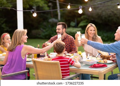 Front View Of A Multi-generation Caucasian Family Outside At A Dinner Table Set For A Meal, Raising Their Glasses And Making A Toast