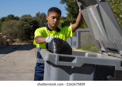 Front view of a municipal worker in uniform throwing a bag of garbage into a large dumpster while holding up the dumpster lid.