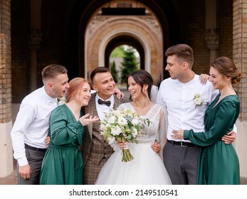 Front view of newlyweds standing between friends, embracing each other and having fun during wedding walk in ancient town