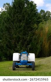 Front view of a nondescript blue tractor in a grassy clearing. Pine forest in the background. Flat Rock, NC 2025