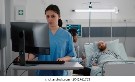 Front View Of Nurse Typing Medical Expertise On Computer While In Background Black Practitioner Doctor Discussing With Sick Man Healthcare Treatment. Hospitalized Patient Having Respiratory Disorder