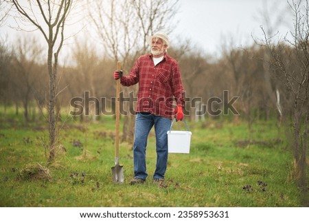 Front view of old man working in orchard in spring. Male gardener in panama hat and gloves standing, looking, holding spade and bucket. Concept of countryside and agriculture.