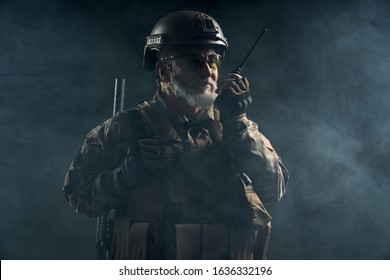 Front View Of Old Military Soldier In Uniform Holding Portable Radio Station In Smoke. Close Up Of Male Commander In Camouflage Uniform Giving Orders In Dark Atmosphere. Concept Of Army, Military.