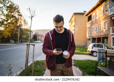 Front View Of One Adult Young Caucasian Man Walking In Front Of Street In Town In Autumn Evening Holding And Using Mobile Phone And Cup Of Coffee Half Length Real People Copy Space Looking To The Side